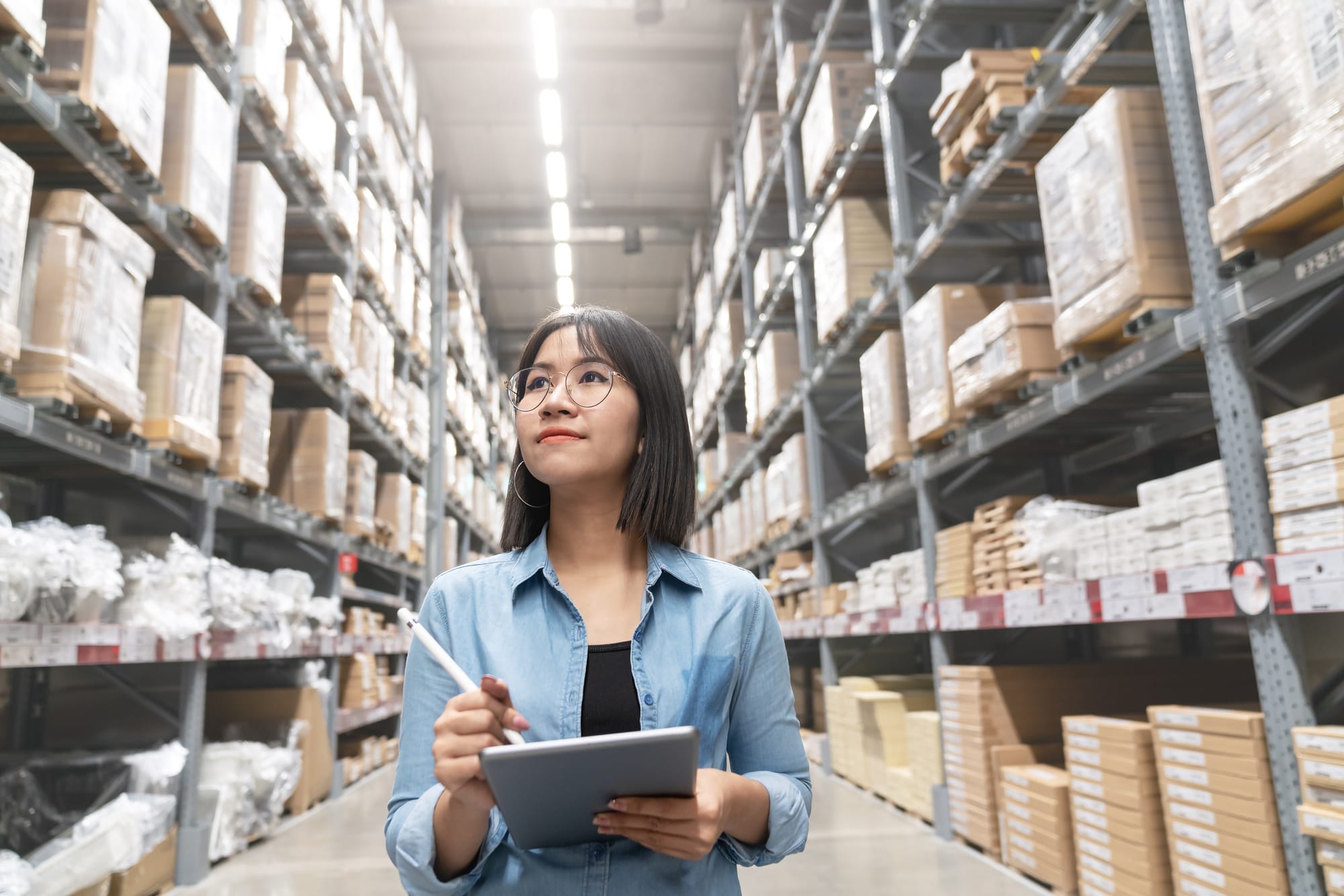 Candid of young Asian woman auditor or trainee staff work looking up stocktaking inventory in warehouse store by computer tablet with wide angle view. Asian owner or small business concept.