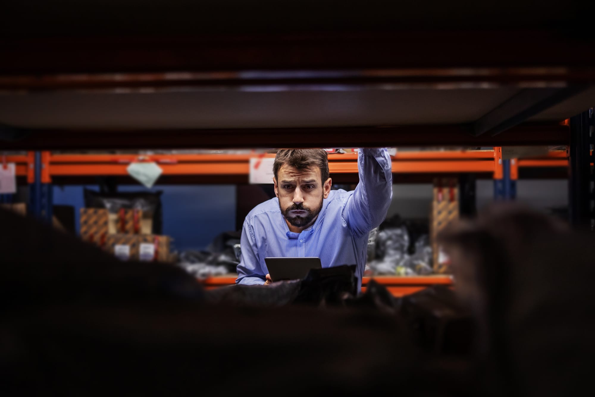 Frustrated supervisor holding tablet and trying to find a box while leaning on shelves in storage