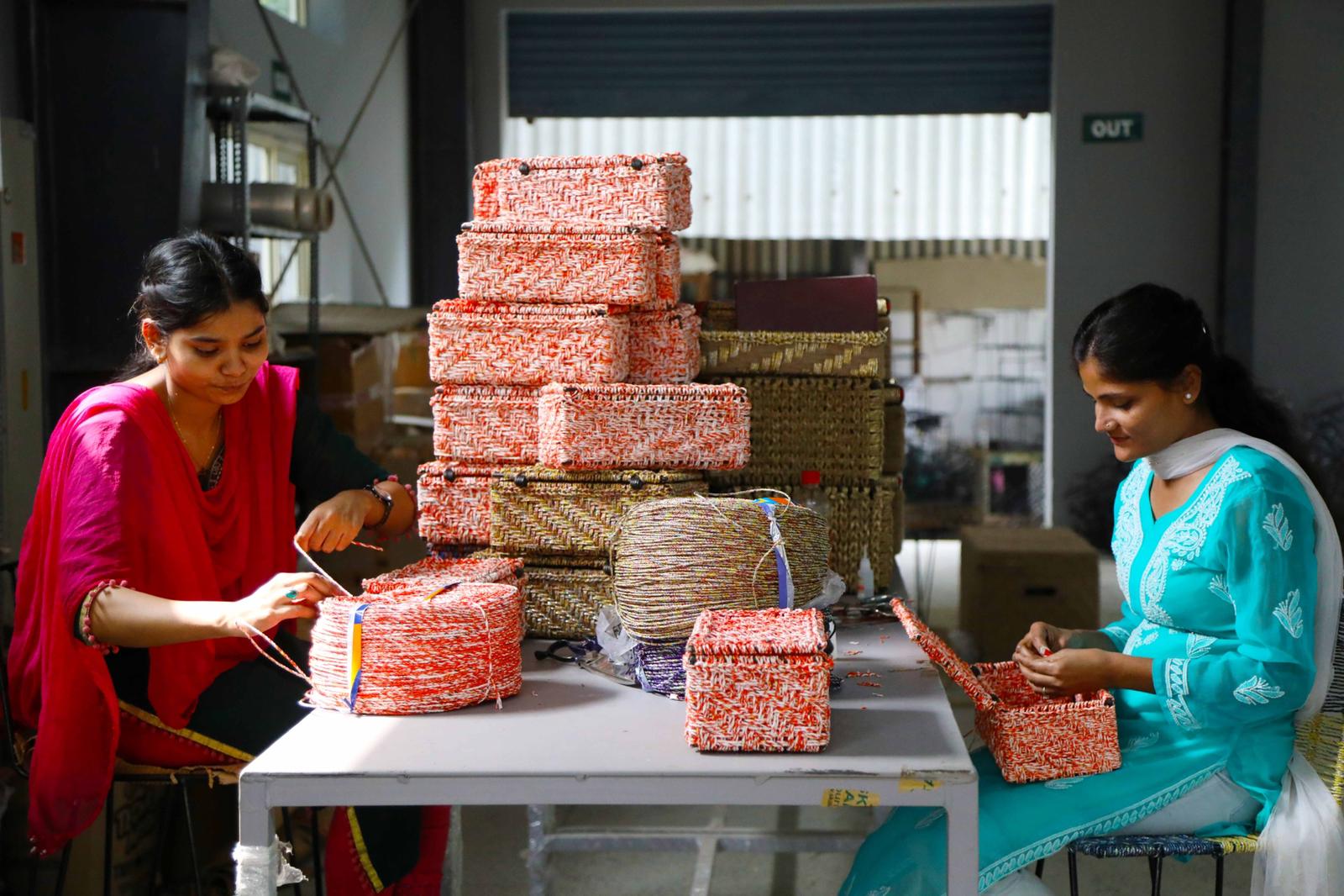 Two women artisans weaving colorful storage baskets by hand, surrounded by stacks of completed pieces.