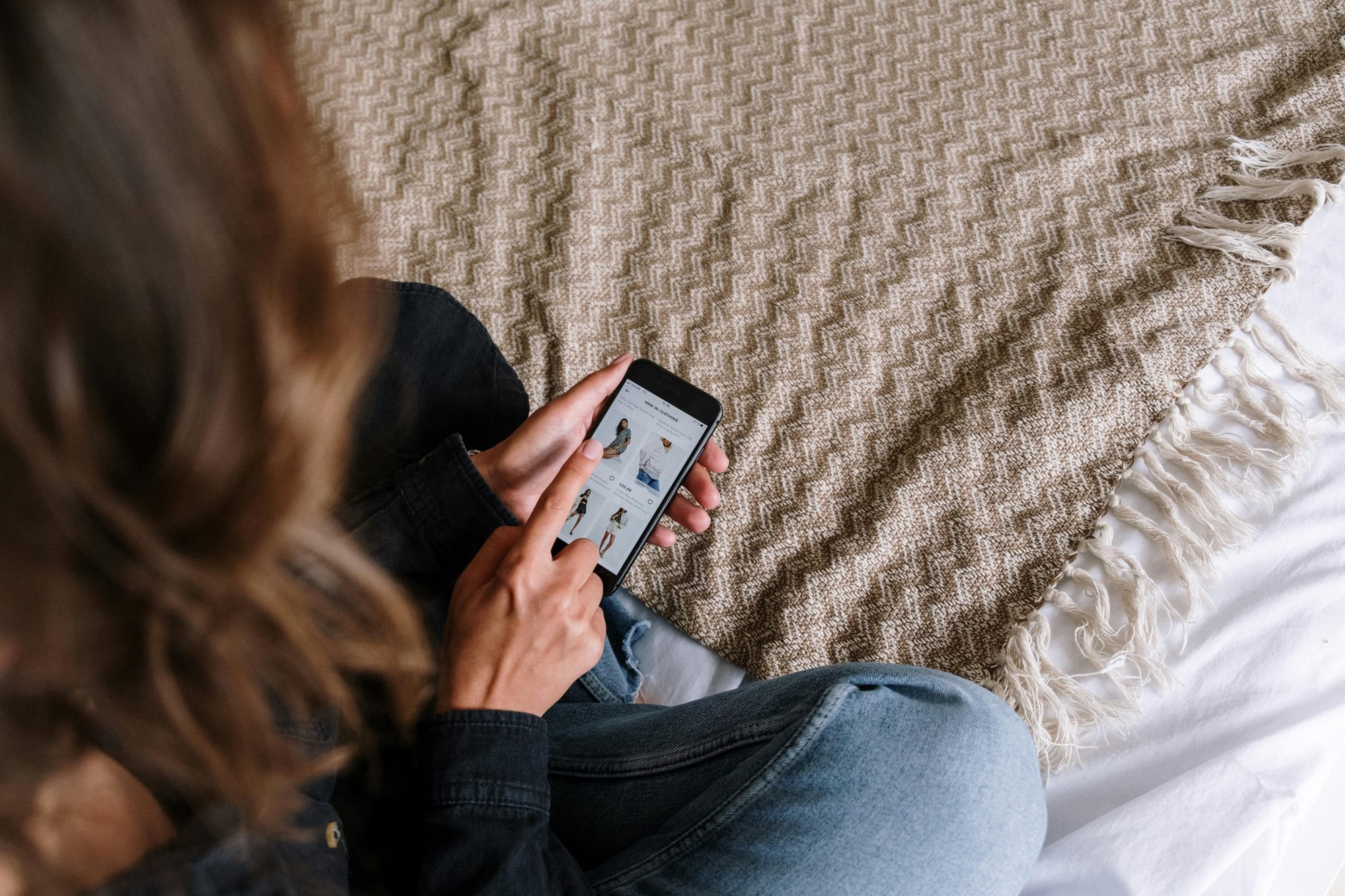 Woman browsing an online store on her phone while sitting on a bed.