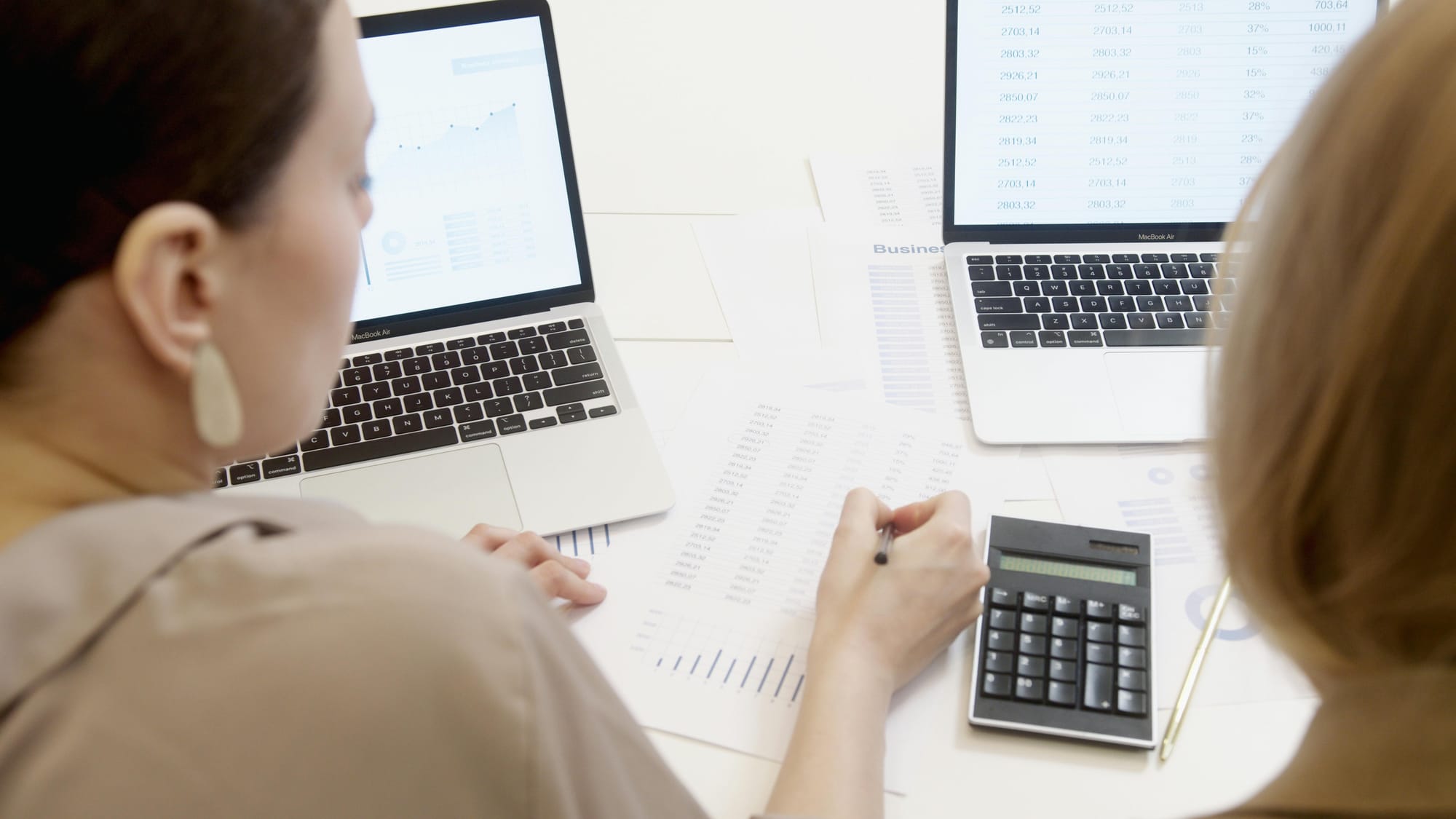 Two professionals analyze inventory and sales data using laptops, printouts, and a calculator during a business meeting.