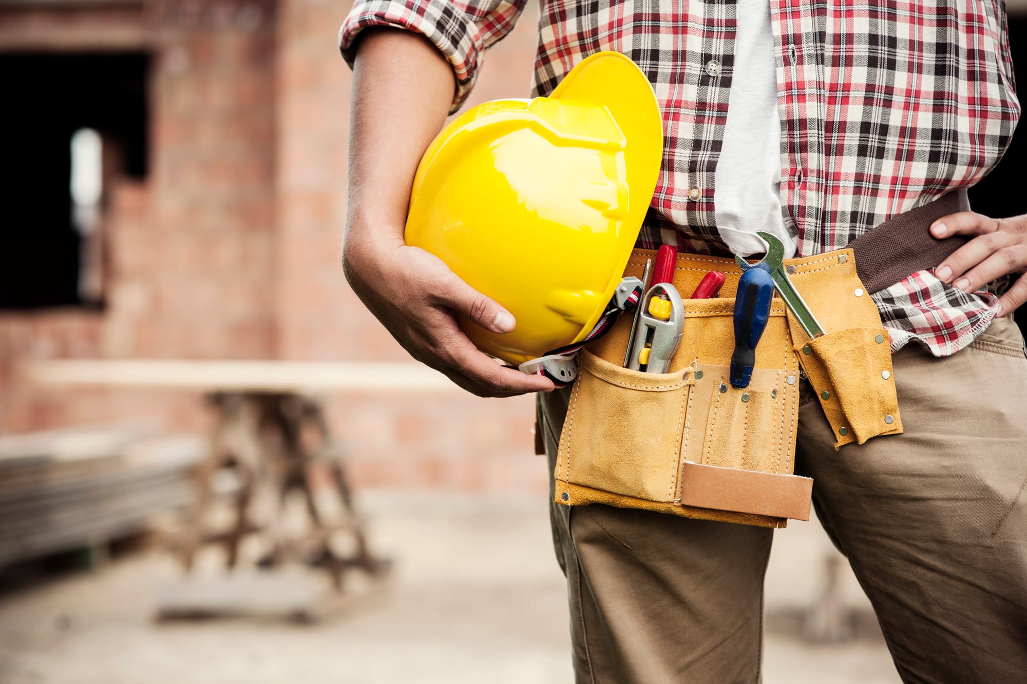 Construction worker holding a yellow hard hat and wearing a tool belt at a building site