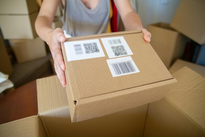 A person hands over a brown package with barcode and QR code labels, surrounded by other shipping boxes.